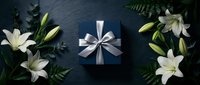 Elegant dark blue gift box with silver ribbon surrounded by white lilies and green foliage on slate background for special occasions and celebrations.