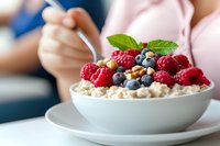 Fresh raspberries and blueberries on top of creamy oatmeal in white ceramic bowl, garnished with mint leaf, healthy breakfast concept with selective focus.