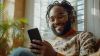Young African American man with dreadlocks and glasses smiling while using smartphone at home, wearing casual sweater near window with natural light and houseplant.