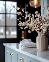 Delicate white cherry blossoms in beige ribbed vase on marble countertop, against dark window backdrop with soft natural lighting and folded linens.