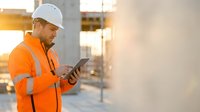 Construction worker in safety gear using tablet at building site during golden hour for project management and digital workflow.