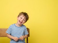 Young boy with light brown hair, Caucasian ethnicity, wearing blue and white striped shirt, laughing joyfully while sitting on wooden chair against bright yellow background.