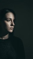 Young Caucasian woman in dramatic side lighting against dark background, contemplative expression, moody portrait photography for editorial use.
