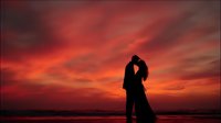 Romantic couple silhouette embracing at sunset beach with dramatic orange red sky clouds creating intimate moment for wedding anniversary celebration.