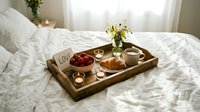 Romantic breakfast in bed tray with croissant, strawberries, coffee, candles, flowers, and a love note on white linen sheets.