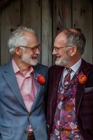 Senior male couple in stylish suits sharing joyful moment at wedding celebration, wearing floral boutonnieres and expressing genuine happiness through warm smiles.