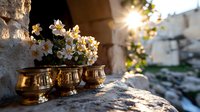 Golden vintage brass pots with white narcissus flowers on stone ledge at sunset, sunlight streaming through ancient stone wall creating magical atmosphere.
