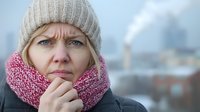 Young Caucasian woman in winter clothing with worried expression, wearing beige knit hat and pink scarf against blurred urban background on cold day.