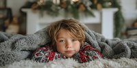 Young child wrapped in cozy gray blanket wearing festive holiday sweater, resting chin on hands with fireplace and Christmas decorations in background.
