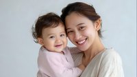 Young Asian mother holding smiling baby girl in pink sweater against white background, expressing joy and tender maternal love in casual lifestyle portrait.