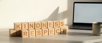 Wooden letter blocks spelling KINDNESS and RESPECT on modern office desk with laptop and coffee mug in warm natural lighting for workplace motivation concepts.