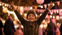 Young boy celebrating with raised arms at festive outdoor event with colorful lights and decorations creating joyful holiday atmosphere.