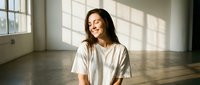Young Asian woman in white t-shirt smiling joyfully in sunlit room with wooden floors and large windows. Natural light creates geometric shadows on wall.