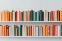 Books in pastel and muted colors arranged on white shelves, creating minimalist composition with orange, pink, green and blue spines in harmonious order.