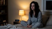 Young woman with dark hair sitting on bed in pajamas looking thoughtful in dimly lit bedroom with warm lamp light creating cozy nighttime atmosphere.