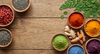 Colorful superfood powders and spices in wooden bowls on rustic table, with goji berries, chia seeds, turmeric, and moringa leaves.