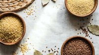 Overhead view of various uncooked grains and seeds in terracotta bowls on light linen fabric with scattered ingredients, suitable for food styling, recipe blogs, and culinary content.