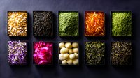 Colorful collection of diverse ingredients in black square bowls on dark background - corn kernels, tea leaves, matcha powder, carrots, herbs, flowers, pickled vegetables.