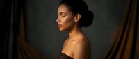 Young African American woman in elegant black dress posing gracefully in dramatic lighting with flowing fabric backdrop for fashion and beauty concepts.