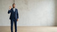 Young male professional in formal navy business suit and tie gesturing okay sign in empty modern office space with concrete wall background.
