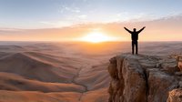 Man celebrating success on mountain cliff edge at sunrise with arms raised overlooking desert valley landscape achieving goals and freedom concept.