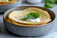 Stack of thin golden crepes with fresh basil leaf and cream filling in gray ceramic bowl, close up view on light background for food blog.