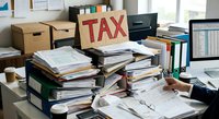 Cluttered office desk with stacks of tax documents, folders and paperwork creating chaotic workspace for accounting and financial planning during busy season.