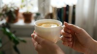 Hands holding warm cup of coffee or tea with latte art in cozy home interior with plants and window light, morning beverage lifestyle.
