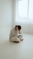 Young Asian woman in beige pajamas sitting cross-legged on white floor holding head in hands showing stress, depression or headache in minimalist room.