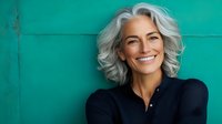 Confident mature woman with silver gray hair and natural smile wearing dark top against turquoise wall background, representing authentic aging and wellness.