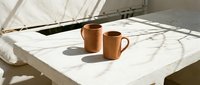 Two terracotta ceramic mugs on white marble table with dramatic window shadows creating minimalist morning coffee scene for kitchen decor and lifestyle branding.