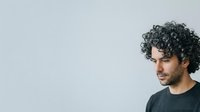 Young man with curly dark hair and olive complexion wearing dark casual shirt, looking downward in contemplative pose against neutral gray background.