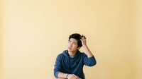 Young Asian male wearing casual blue denim shirt against warm yellow background, looking upward thoughtfully while touching head, suitable for lifestyle, education, and corporate design projects.
