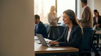 Young woman of South Asian descent wearing professional business attire and headset, working on laptop at desk in modern office environment with colleagues collaborating in background during business meeting.