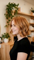 Happy young Caucasian woman with wavy bob haircut smiling in profile wearing black top in modern kitchen with wooden shelves and green plants.