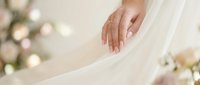 Elegant bride hand with gold engagement ring touching soft white wedding dress fabric, floral bokeh background, romantic bridal detail.