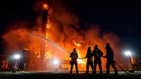 Firefighters battle massive industrial blaze with water hoses as flames and smoke engulf building structure during emergency response operation at night.