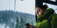 Young woman in green winter jacket checking smartphone while traveling by train, snowy forest landscape visible through window. Peaceful winter journey.