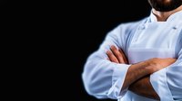 Professional male chef in white uniform with crossed arms against dark background, dramatic lighting highlights confidence and expertise in culinary field.