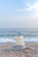 Stacked sea glass and smooth stones balanced on sandy beach with ocean waves and soft sky in background, representing zen, mindfulness and coastal relaxation.