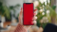 Female hand with red manicure holding smartphone with blank red screen against blurred green plant background, perfect for app mockup.