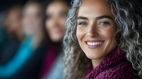 Confident mature woman with natural gray curly hair and bright smile wearing purple knitted scarf, looking at camera. Blurred people in background.
