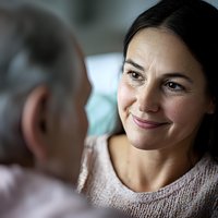 Middle-aged Asian woman with gentle smile wearing knit sweater looks lovingly at elderly person during caring conversation in soft natural lighting.