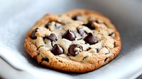Freshly baked chocolate chip cookie on white plate with golden brown edges and melted chips, ready for snacking or dessert photography.