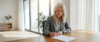 Professional blonde businesswoman in gray suit signing contract documents at modern office desk with natural lighting and plants in background.