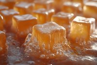 Soft caramel candy cubes with melted drips arranged in rows on reflective surface, macro shot showing rich texture and golden amber color in warm lighting.