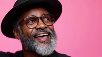 Cheerful mature African American man with gray beard wearing glasses and black hat laughing against pink background, expressing genuine joy and happiness.