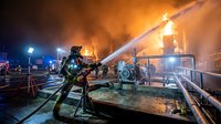 Firefighter in protective gear battles intense industrial blaze with water hose as emergency crews respond to dangerous warehouse fire at night.