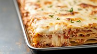 Classic homemade lasagna with layers of pasta, ground meat sauce, melted mozzarella and ricotta cheese garnished with fresh herbs on metal baking pan.