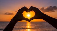 Hands forming heart shape silhouette against golden sunset over ocean water. Romantic love symbol gesture at beach during twilight hour for Valentine concept.